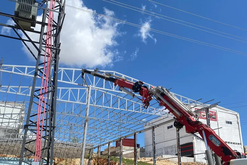Vinçli kamyon, çelik çatı makasını yapının üzerine yerleştiriyor; solda trafo direği ve enerji hattı duruyor.