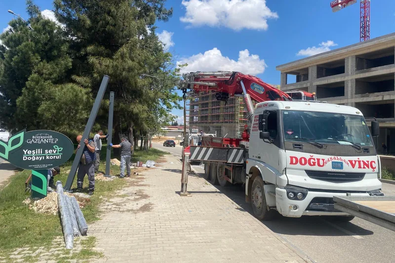 Kaldırım kenarında dikilen çelik direk; yerde sırasını bekleyen ikinci direkler ve arkada vinçli kamyon.